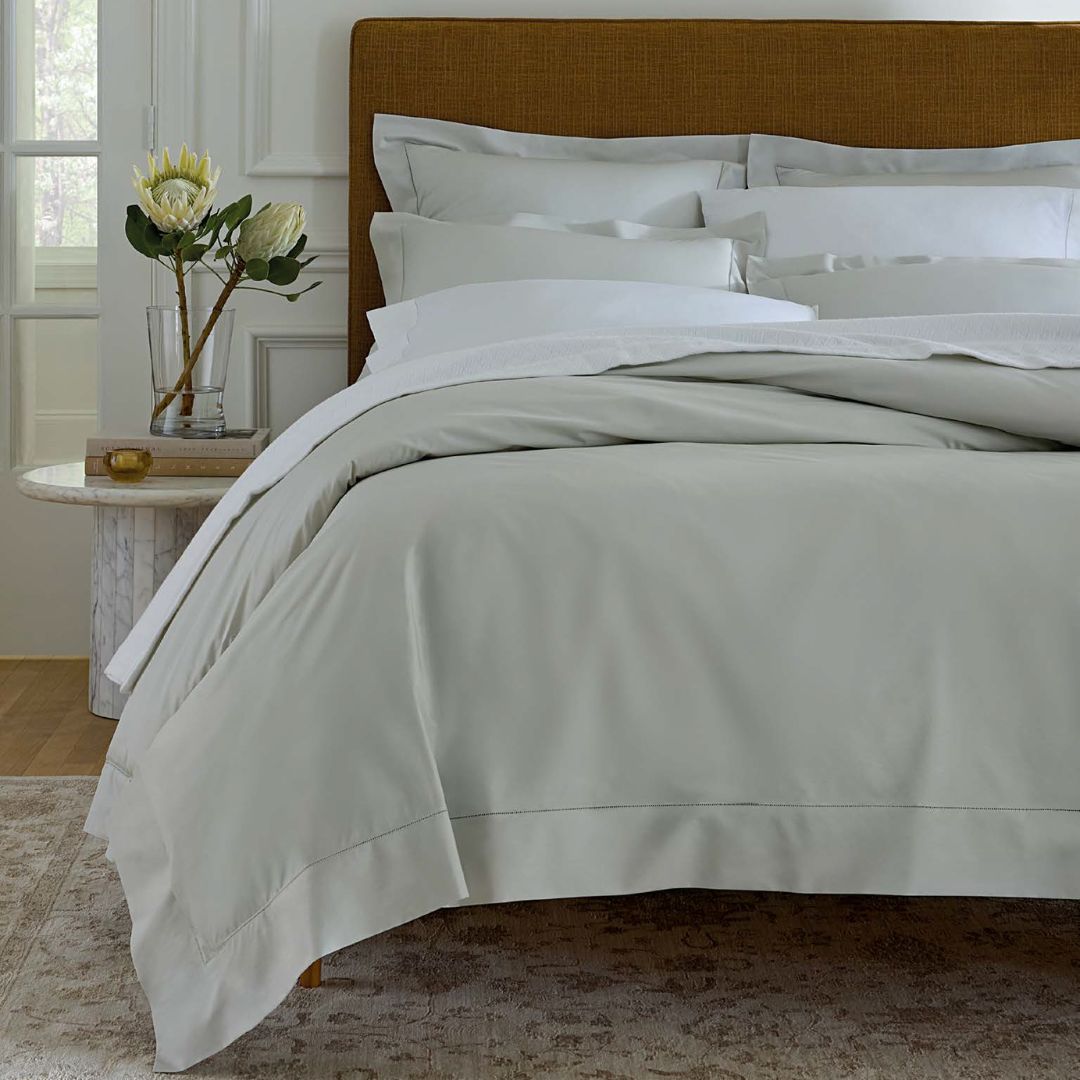 A neatly made bed with light gray Sferra Luxor Bedding beside a round table holding a vase of flowers and books.