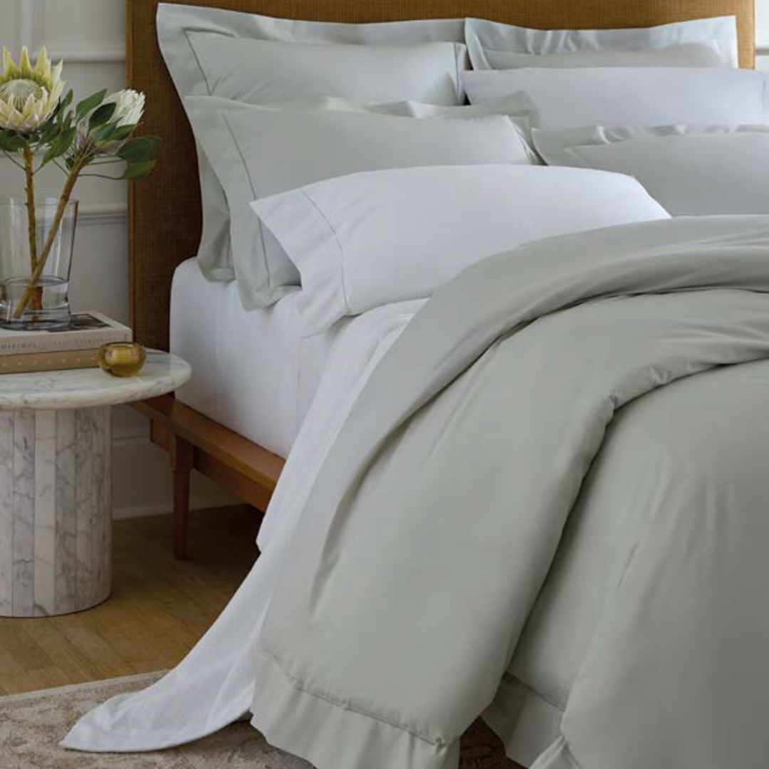 A neatly made bed with Sferra Luxor Bedding next to a marble side table holding books and a vase of flowers.