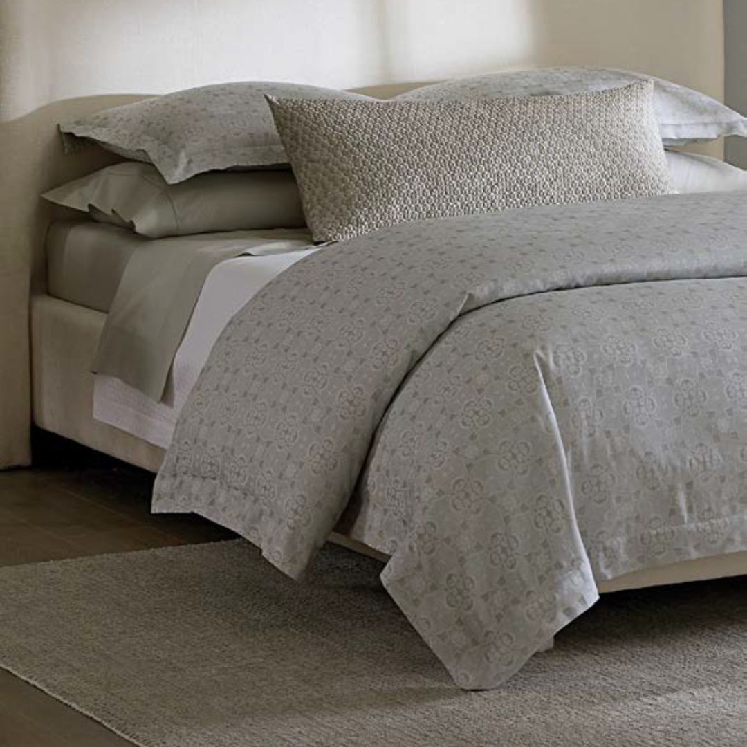 A neatly made bed with luxury bedding, stacked pillows, and a decorative cushion on a light-colored rug.
