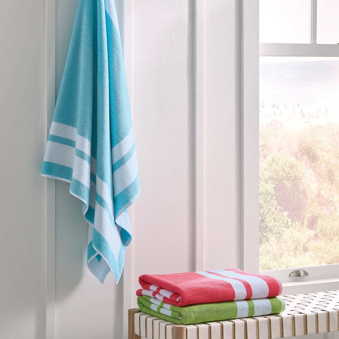 A 100% cotton beach towel hangs on a white wall; luxury beach towels are folded on a bench by a window.