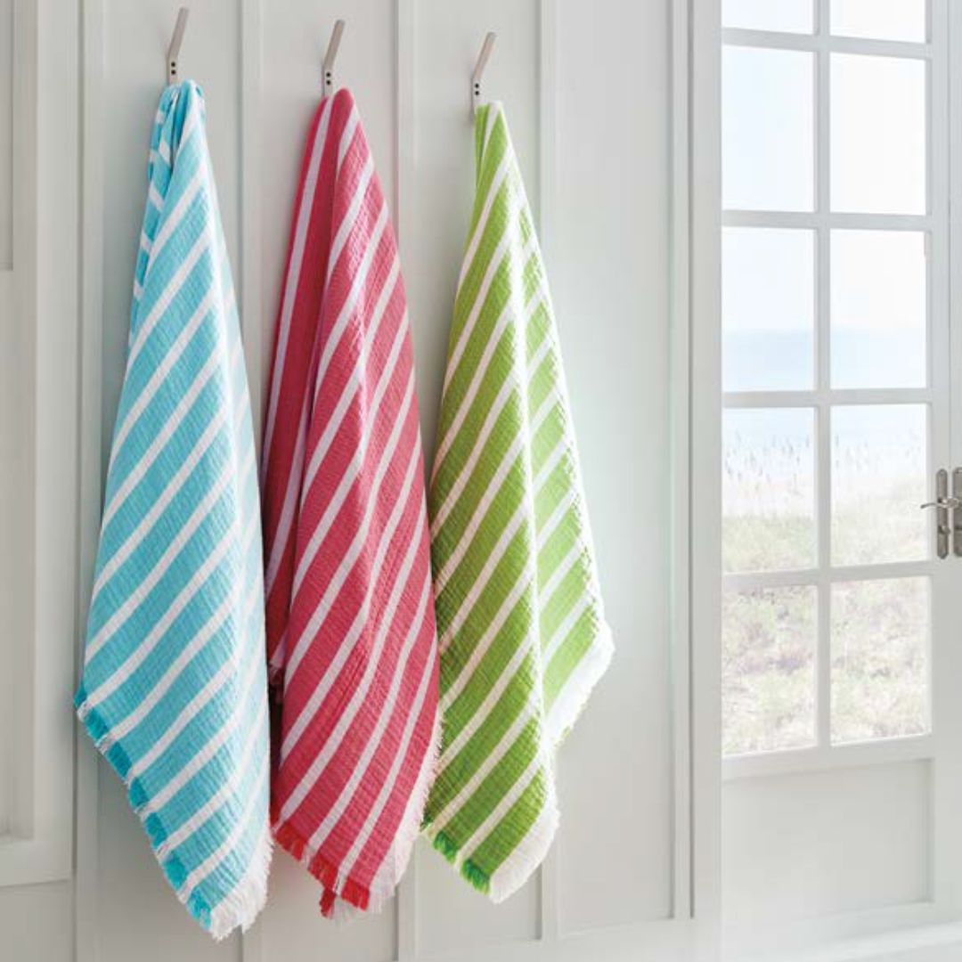 Three striped towels—blue, red, and green—hang on hooks, each a luxury cotton beach towel by the sunny window.