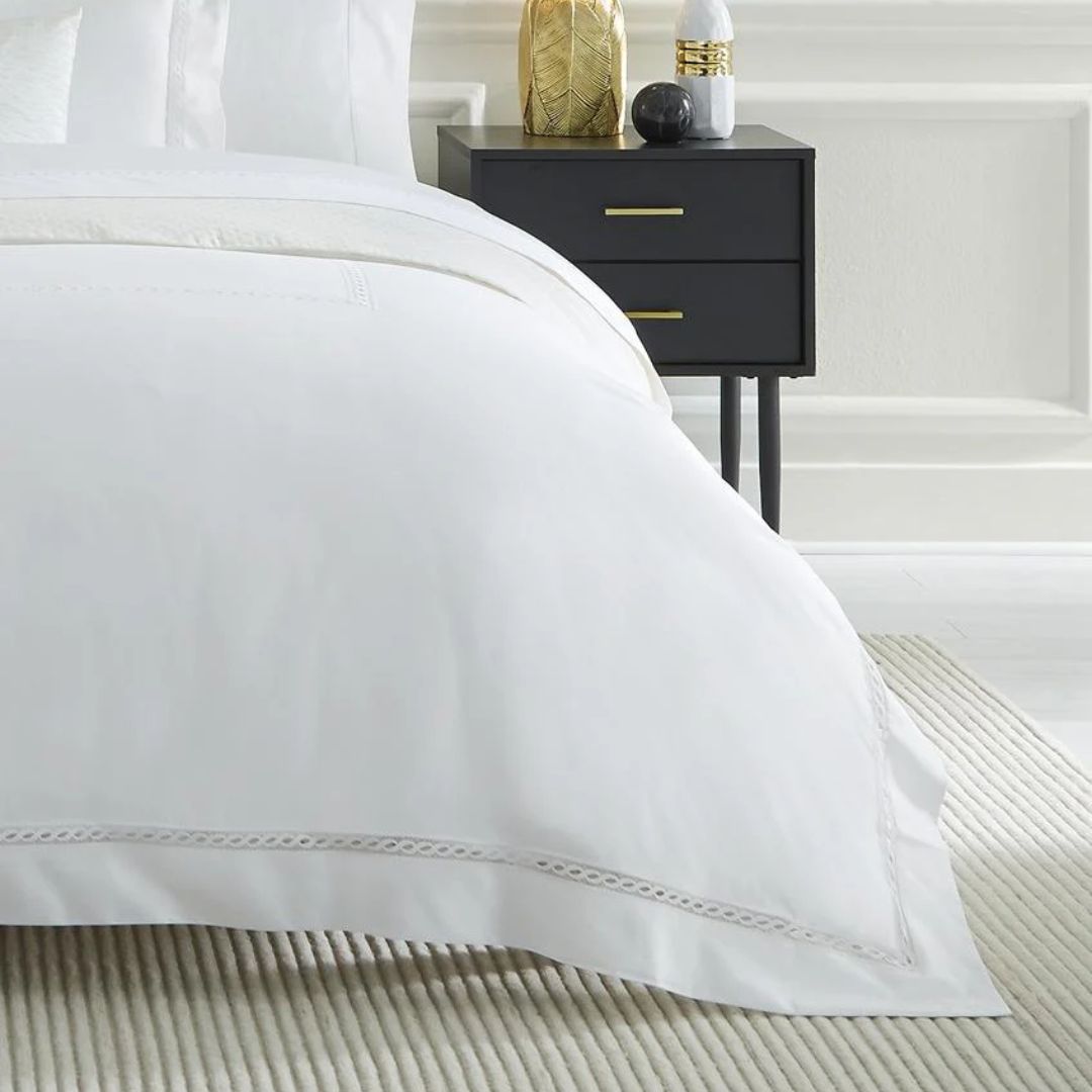 White bedding with a decorative edge—luxury Egyptian cotton sheets—on a bed beside a black nightstand and striped rug.