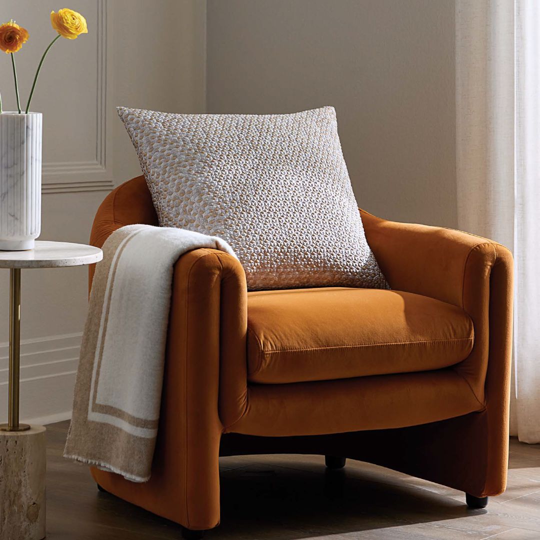 A rust-colored armchair with mineral-inspired texture and a Sferra Nesso Decorative Pillow, beside a vase of yellow flowers.