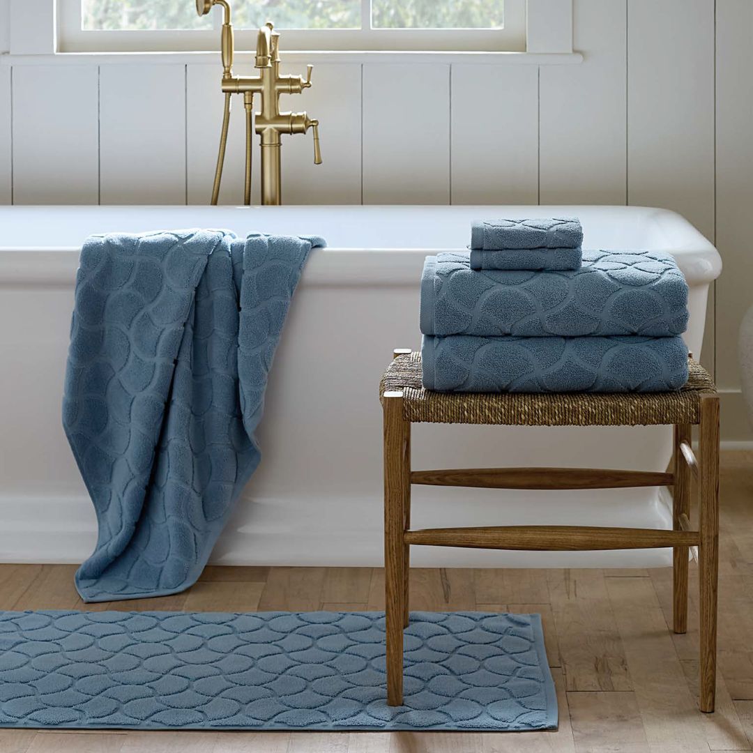 A white bathtub with a blue Sferra Oasi Bath Towel draped over it and luxury bath towels stacked on a nearby chair.
