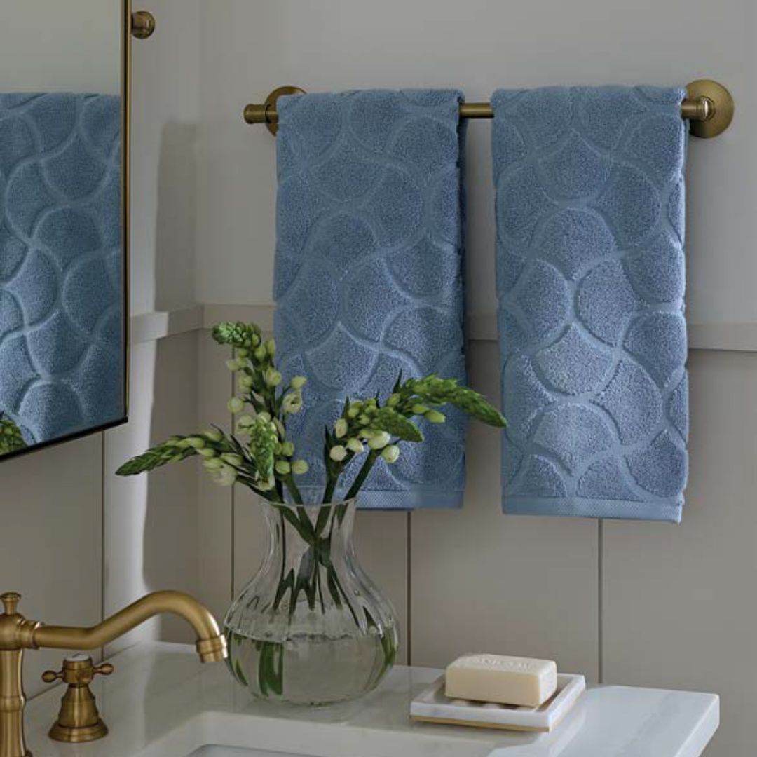 Two blue luxury bath towels hang on a gold towel bar above a sink with soap and a glass vase of white flowers.