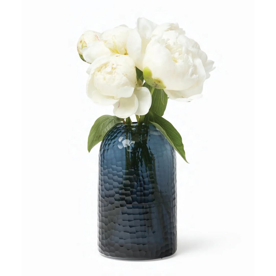 White peonies with green leaves in a polished glass vase, set against a white background.