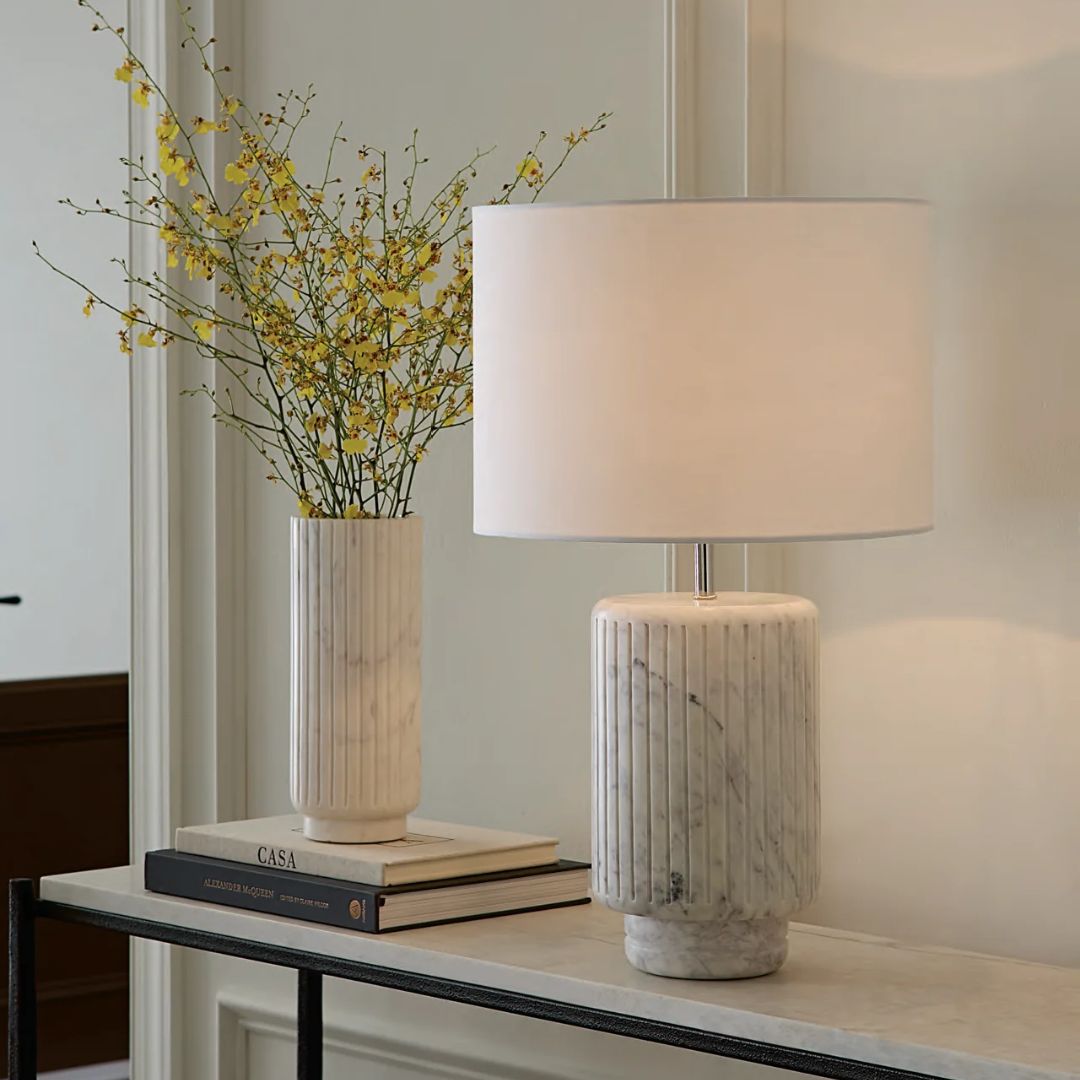 A Sferra Romano Marble lamp and vase with yellow flowers grace a console table with books, against a beige wall.