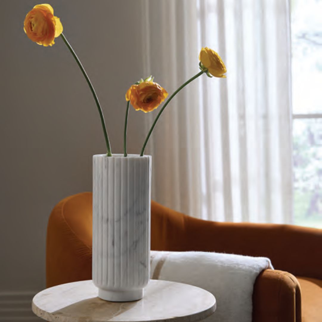 White Sferra Romano Marble vase with orange flowers sits on a round table by an orange chair and white curtains.