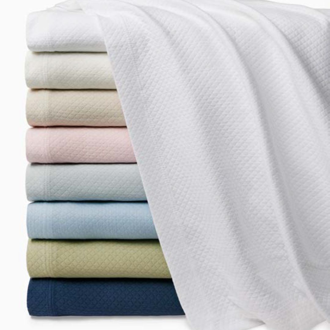 A stack of folded blankets in various colors, topped with a white Rombo coverlet draped over the top.