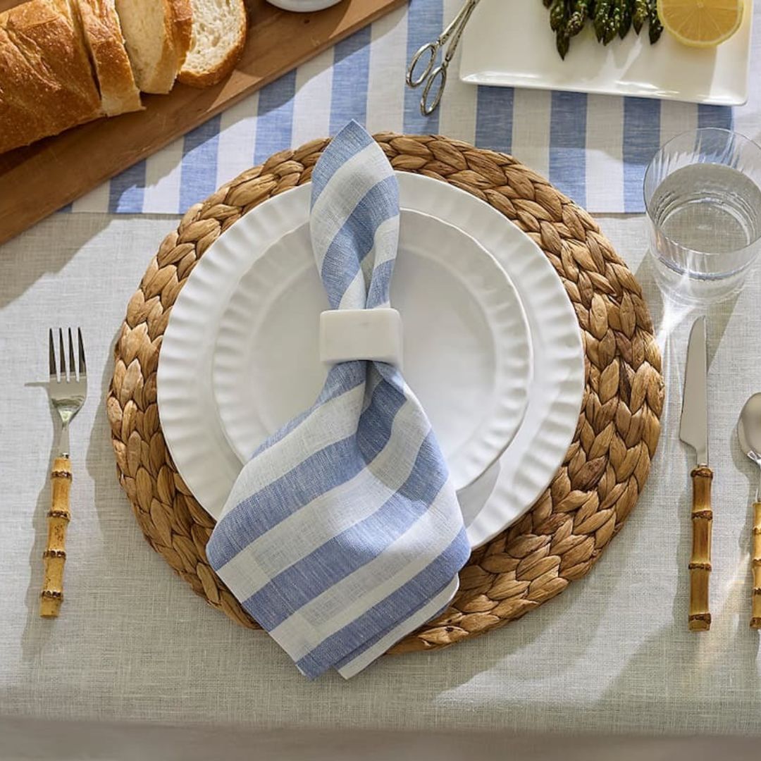 A place setting with coastal table decor—woven placemat, white plates, blue-striped napkin, and bamboo-handled cutlery.