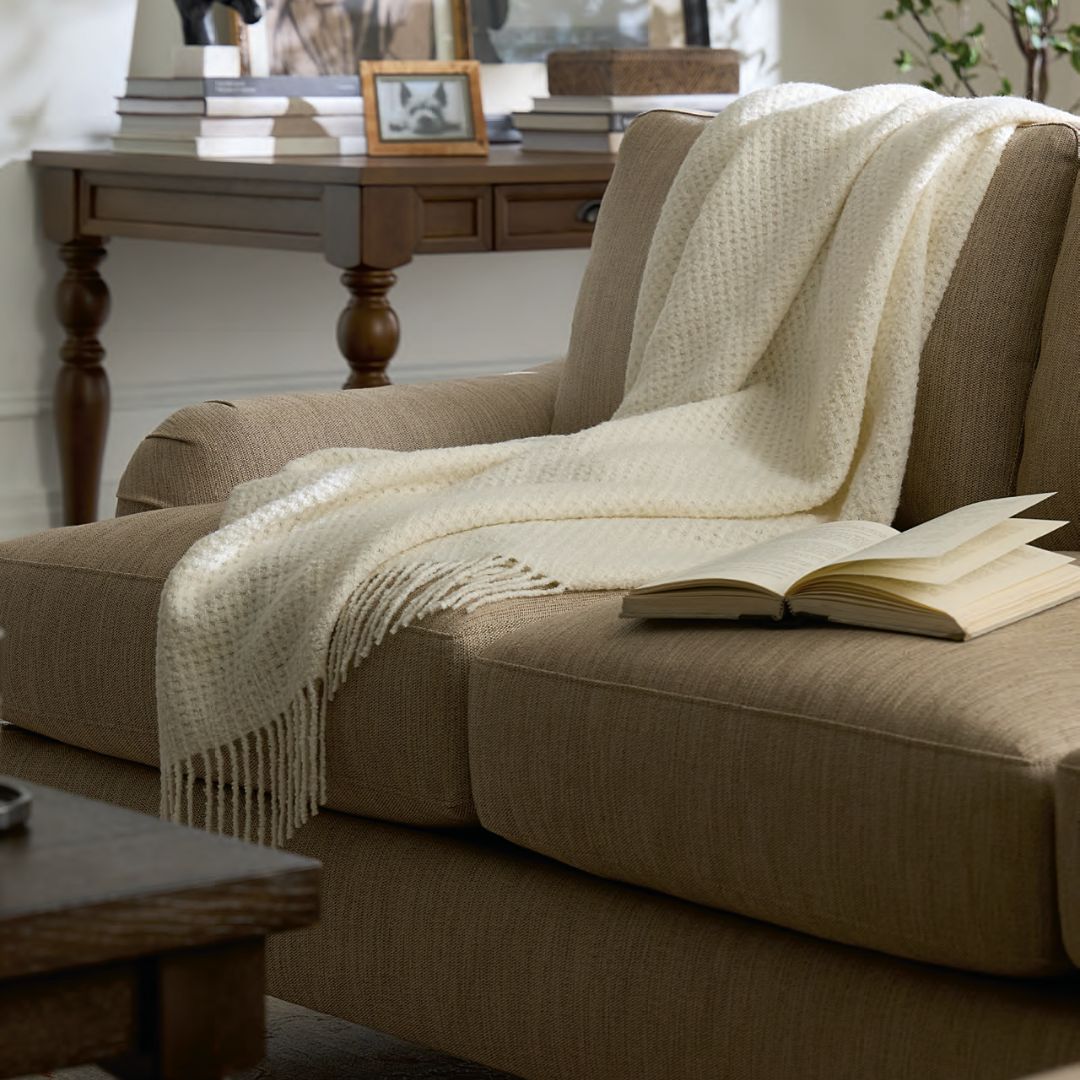 A beige sofa with a Sferra Terranova Throw and an open book; a table and framed photos in the background.