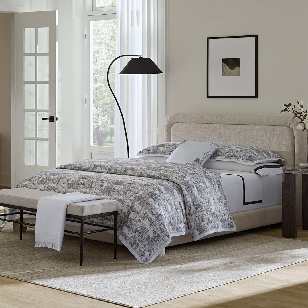 Neatly made bed with floral bedding in a bright bedroom with large windows.