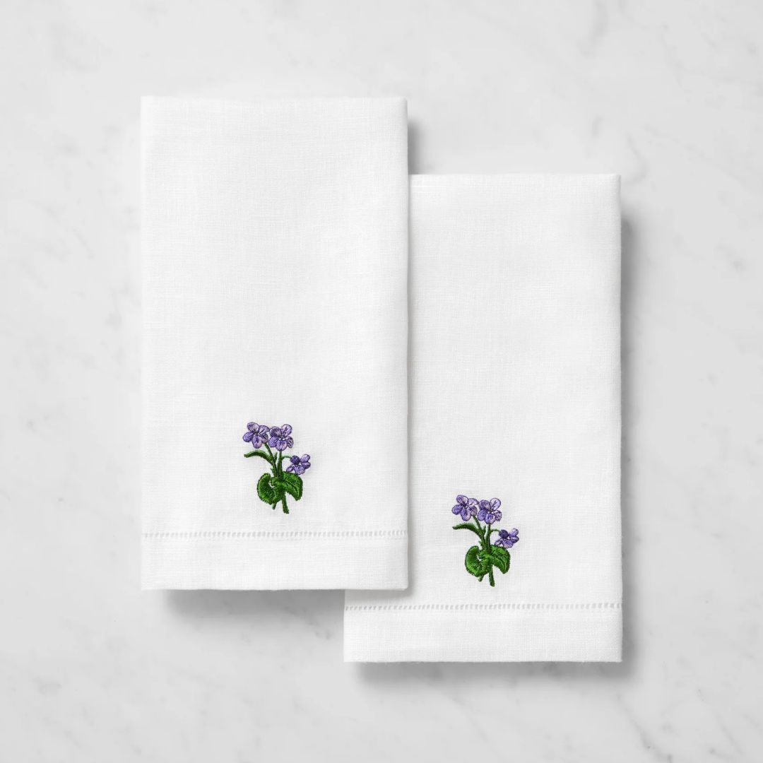 Two white embroidered linen guest towels with purple flowers and green leaves rest on a marble surface.