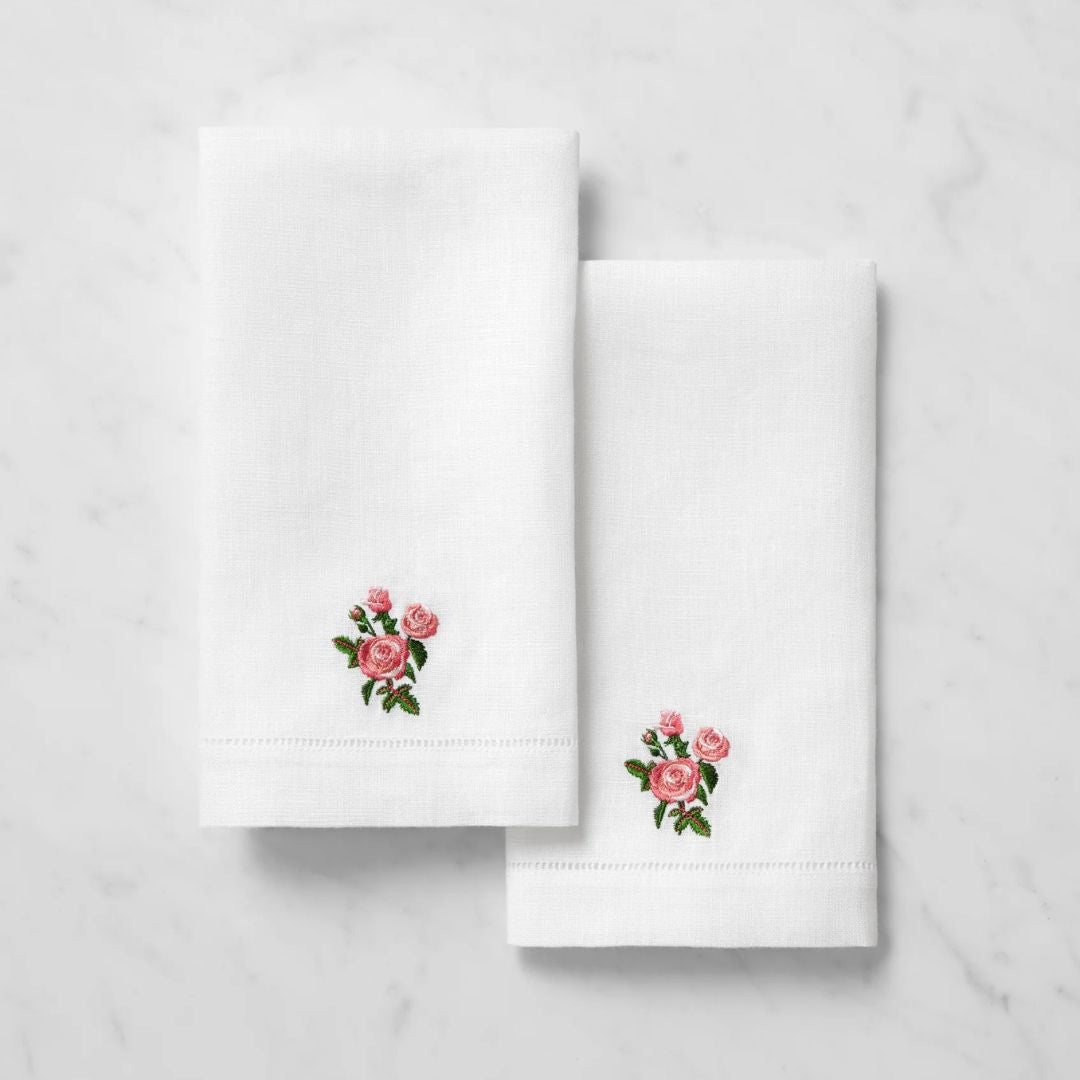 Two white linen guest towels with embroidered pink roses and green leaves, displayed on a white marble surface.