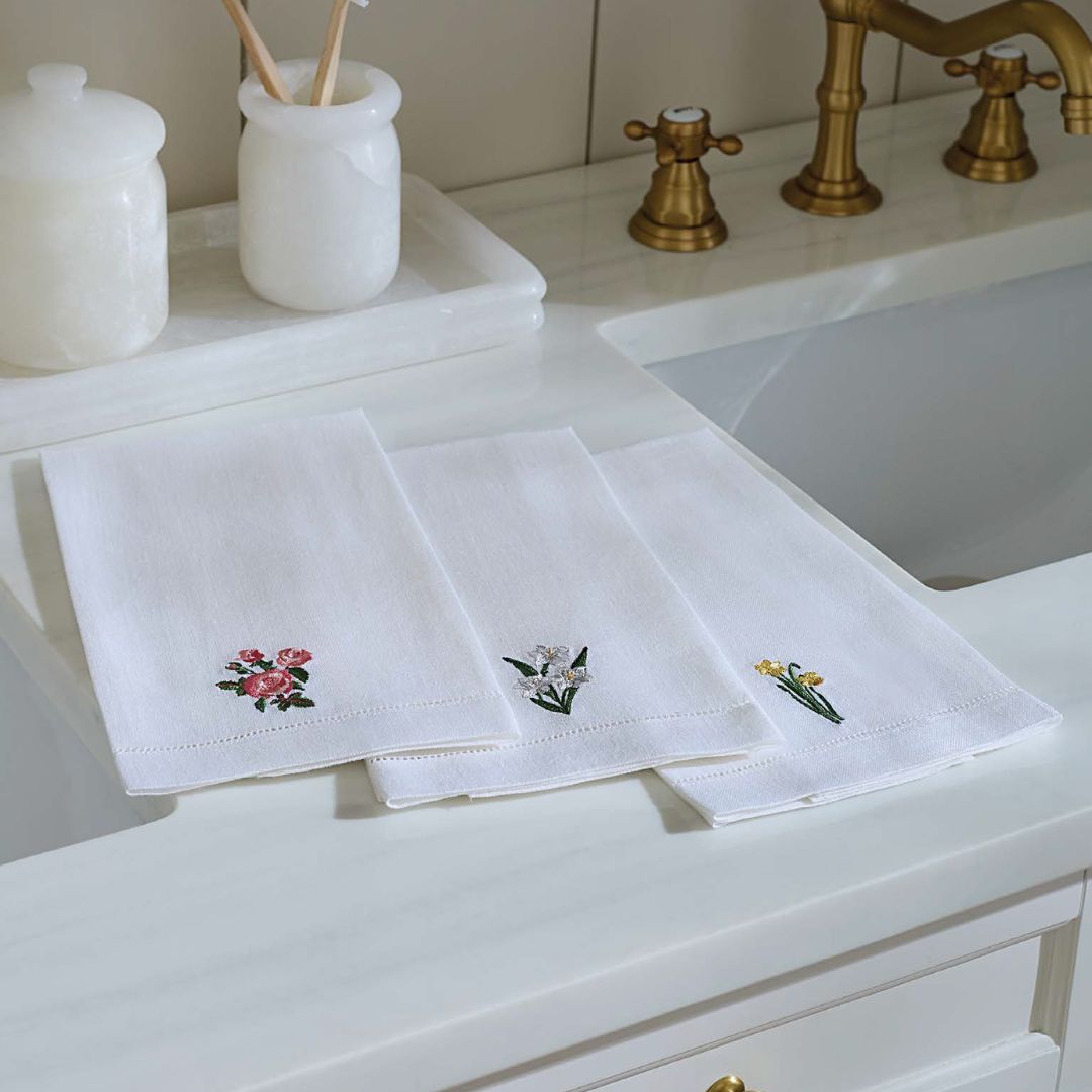 Three white embroidered guest towels with floral designs are laid out on a marble countertop next to gold sink fixtures.