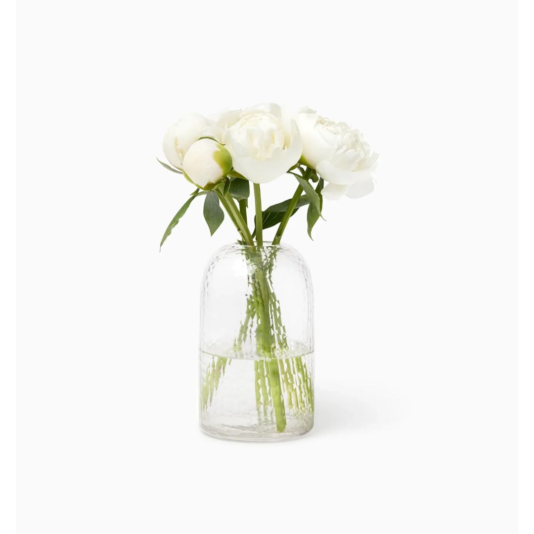 A polished glass vase holds white peonies with green stems and leaves, set against a plain white background.