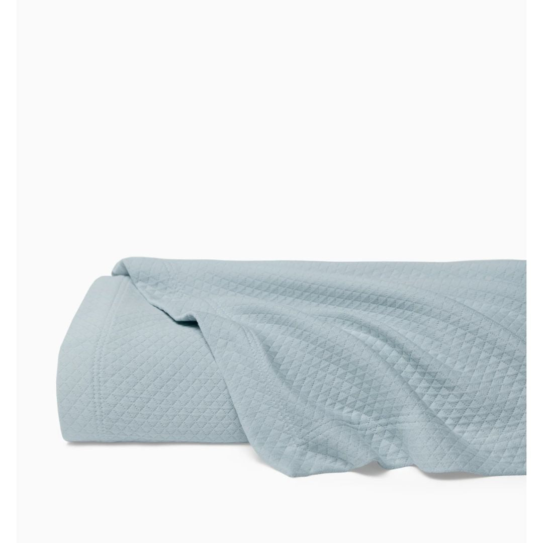 A light blue cotton matelassé blanket is spread out and partially folded on a flat surface against a white background.