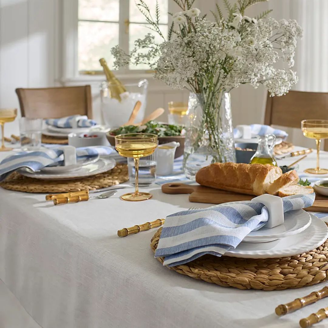 A dining table set with plates, linen table linens, striped napkins, bread, flowers in a vase, and gold-colored drinks.