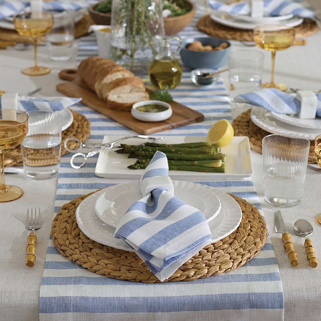 A coastal table decor scene with striped napkins, white plates, bread, asparagus, and woven placemats on a striped runner.