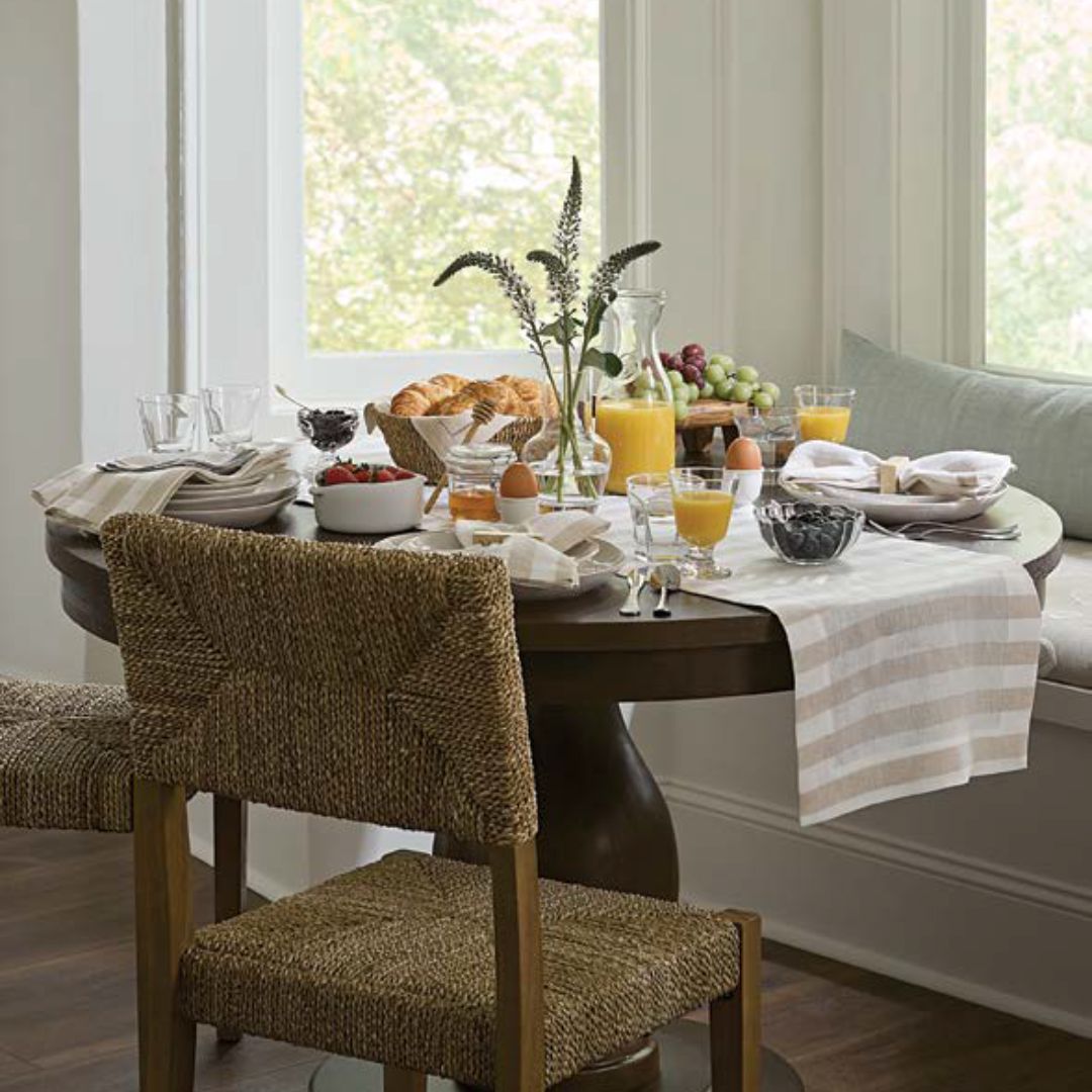 A round table set for breakfast with eggs, croissants, juice, fruit, and coastal table decor by a window.
