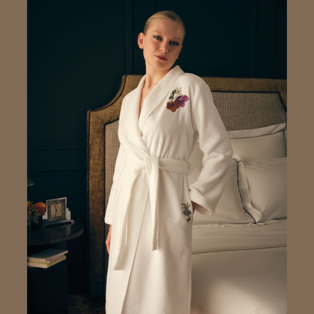 Woman stands by a bed in a white robe with floral embroidery, Yves Delorme Fleurs Bath Collection towels nearby.