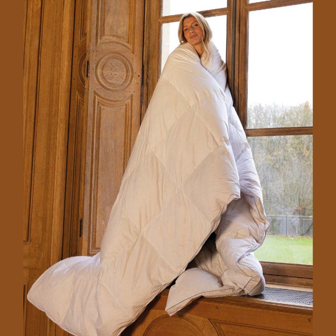 A person wrapped in a down comforter stands by a tall wooden window, gazing outside at the world beyond.