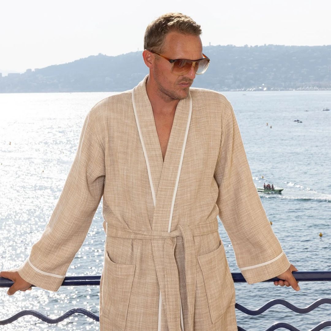 A man in a lightweight honeycomb cotton bathrobe and sunglasses leans against a railing overlooking the sea.