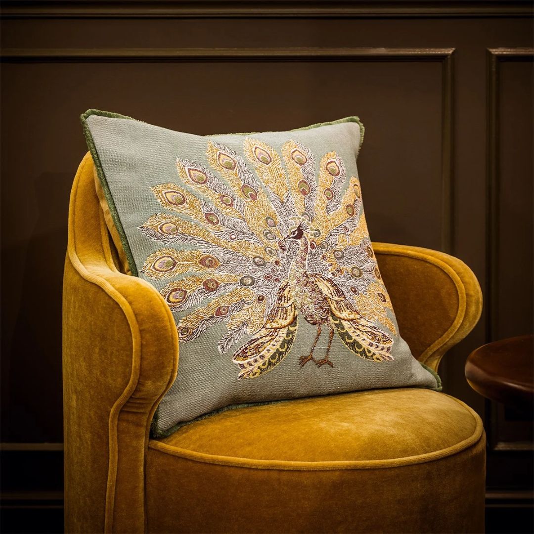 Yves Delorme decorative pillow featuring an embroidered peacock with a fanned tail, resting on a yellow armchair.