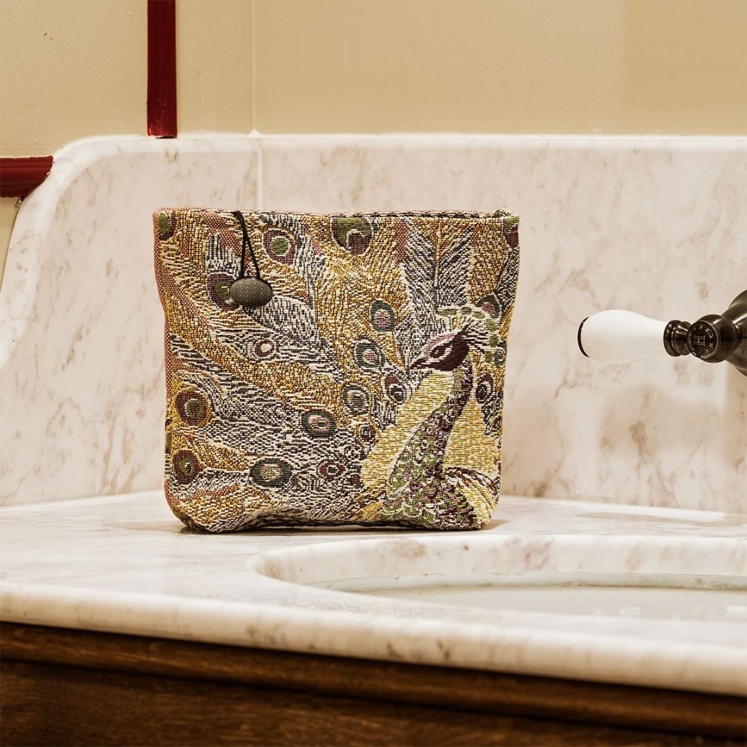 Small, square Yves Delorme tapestry pouch featuring a colorful peacock design on a marble bathroom vanity.