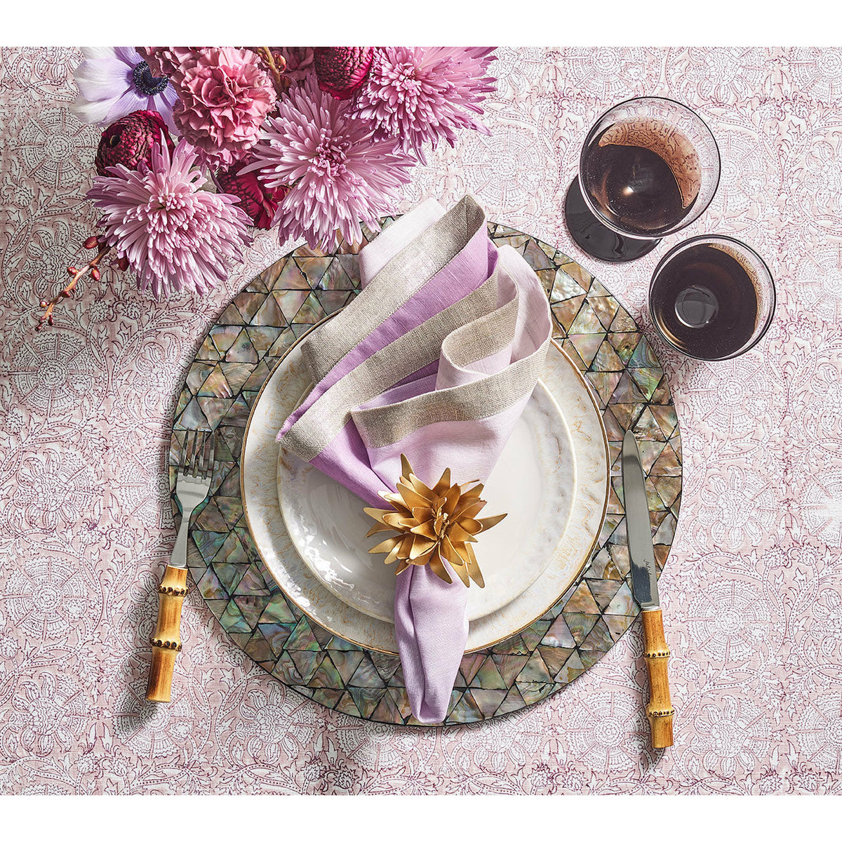 Table setting with Kim Seybert floral napkin rings, plates, wine glasses, cutlery, and pink flowers on lace tablecloth.