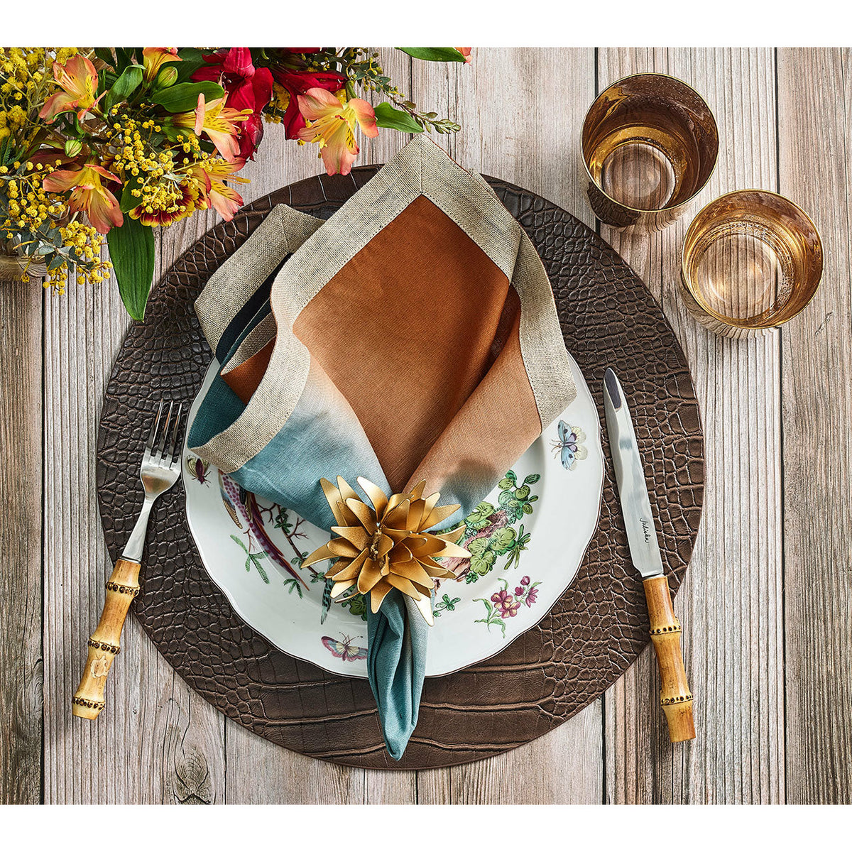 Table setting with Kim Seybert gold napkin ring, floral plate, knife, fork, two glasses, and a bouquet of flowers.