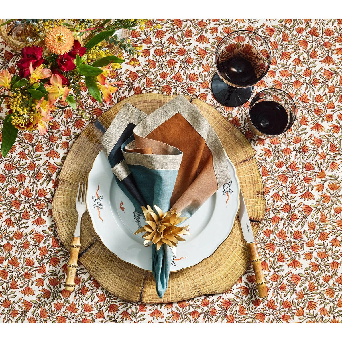 A place setting with Kim Seybert gold napkin rings, two glasses of red wine, and flowers on a leafy patterned tablecloth.